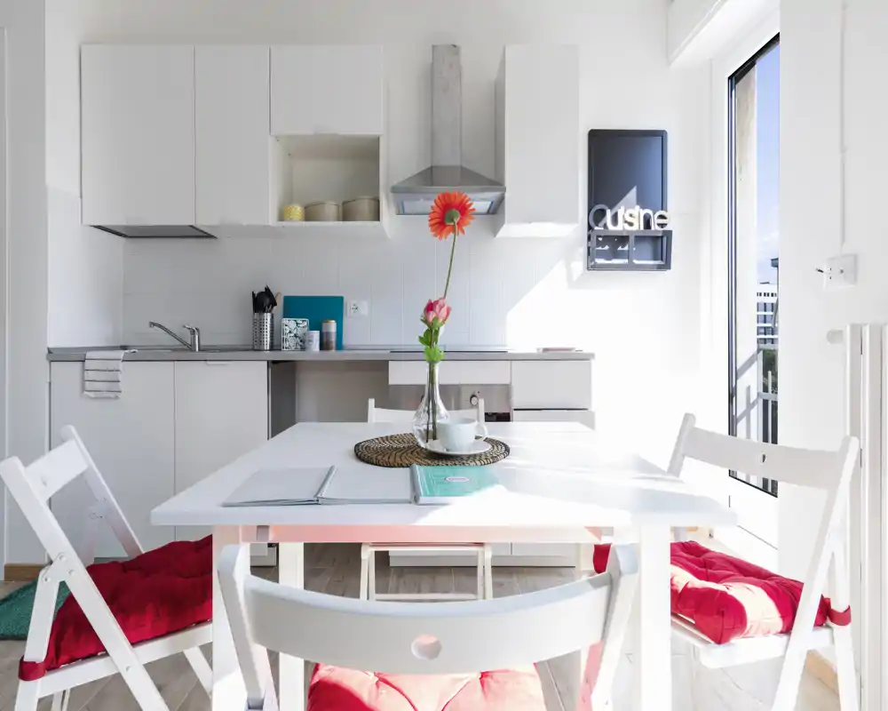 Bright, minimalist open-plan kitchen with a white dining table, red seat cushions and a vase with fresh flowers; large window/door provides abundant natural light.