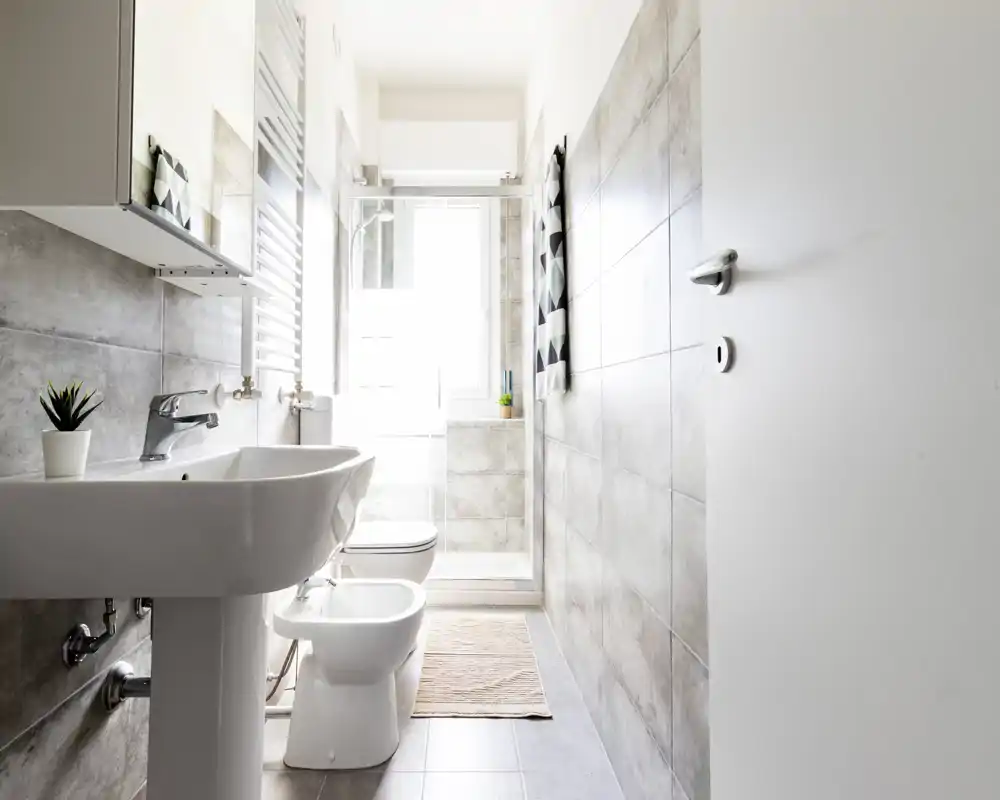 Bright modern bathroom with pedestal sink, bidet, toilet and a glass shower at the far end; neutral grey tiles and natural light create a fresh, clean atmosphere.