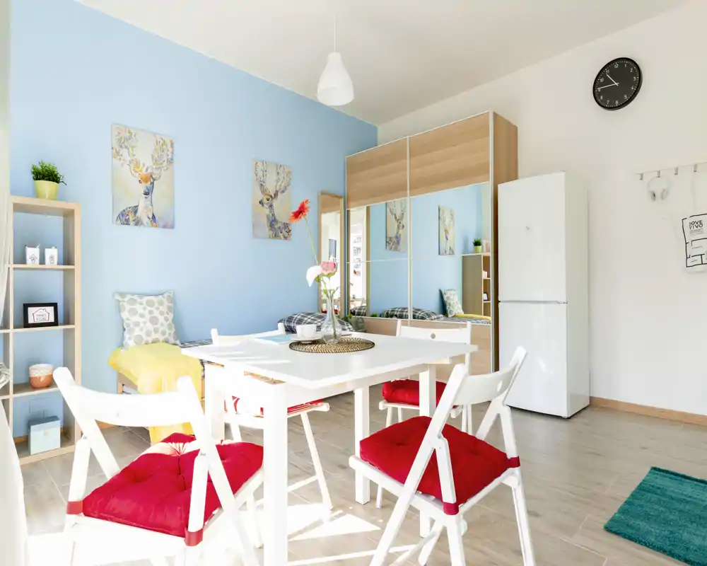 Bright, modern living/dining area with a white table and red-cushioned chairs, light wood floors, decorative wall art and a mirrored wardrobe that increases the sense of space.