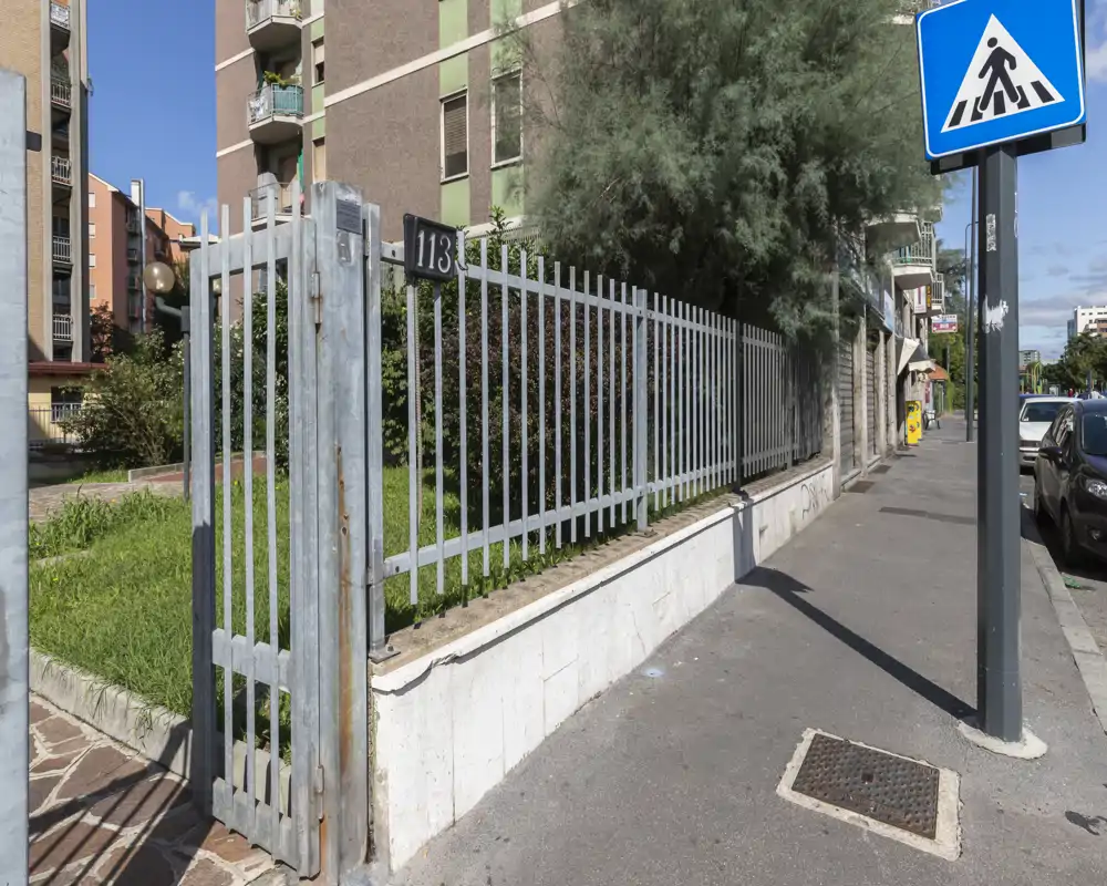Street-side outdoor view showing a metal fence and gate in front of a residential building, with sidewalk, parked cars and a pedestrian crossing sign.