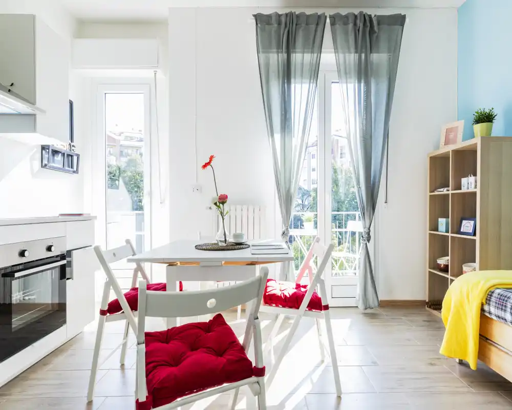 Bright, modern bedroom corner with a single bed at the right, a small dining table with red-cushioned chairs, and a kitchenette by the left wall. Large windows with sheer curtains flood the space with natural light, creating an airy, welcoming ambiance.