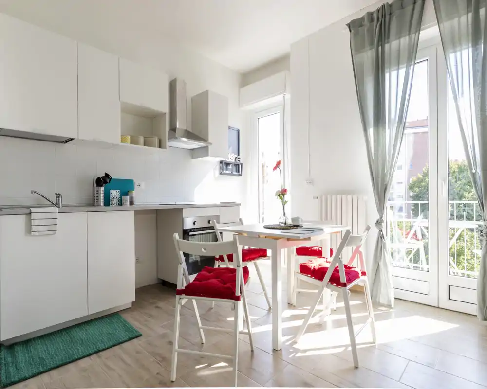 Bright modern kitchenette with white cabinets, a small dining table and red-cushioned chairs beside a sunlit balcony door, ideal for a cosy urban studio.