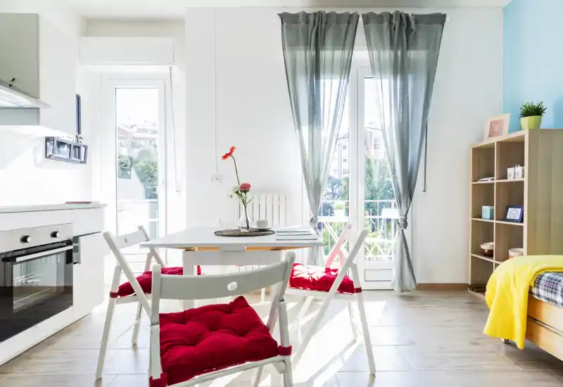 Bright, modern bedroom corner with a single bed at the right, a small dining table with red-cushioned chairs, and a kitchenette by the left wall. Large windows with sheer curtains flood the space with natural light, creating an airy, welcoming ambiance.