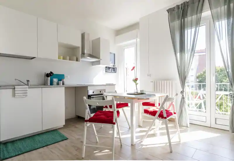 Angolo cottura moderno e luminoso con mobili bianchi, un piccolo tavolo da pranzo e sedie con cuscini rossi vicino alla porta del balcone, perfetto per un monolocale accogliente.