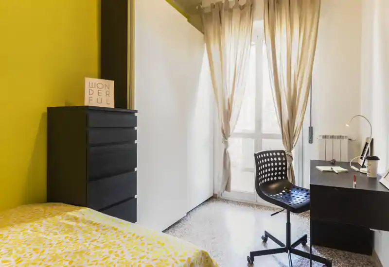 Sunny, neatly staged bedroom with a yellow-accented wall, patterned bedding and a workspace by the window. Large wardrobe and chest of drawers provide storage while sheer curtains let in soft natural light.