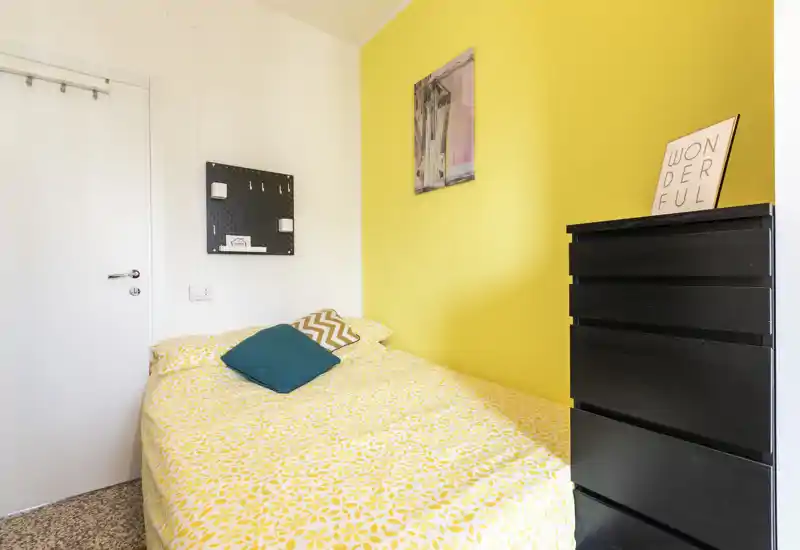 Compact, cheerful bedroom with a yellow accent wall, patterned yellow bedspread and decorative cushions. A tall black dresser and wall decor complete the tidy layout, perfect for a single occupant.