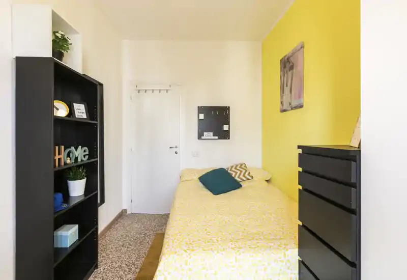 Sunny, compact bedroom with a full single bed dressed in yellow patterned linens, decorative cushions and contrasting black storage furniture — bright, fresh and well styled for a rental listing.