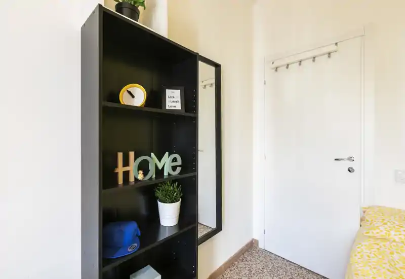 Small bedroom corner with a black shelving unit, decorative items and a partial view of the bed with yellow patterned bedding; bright, tidy and modern.