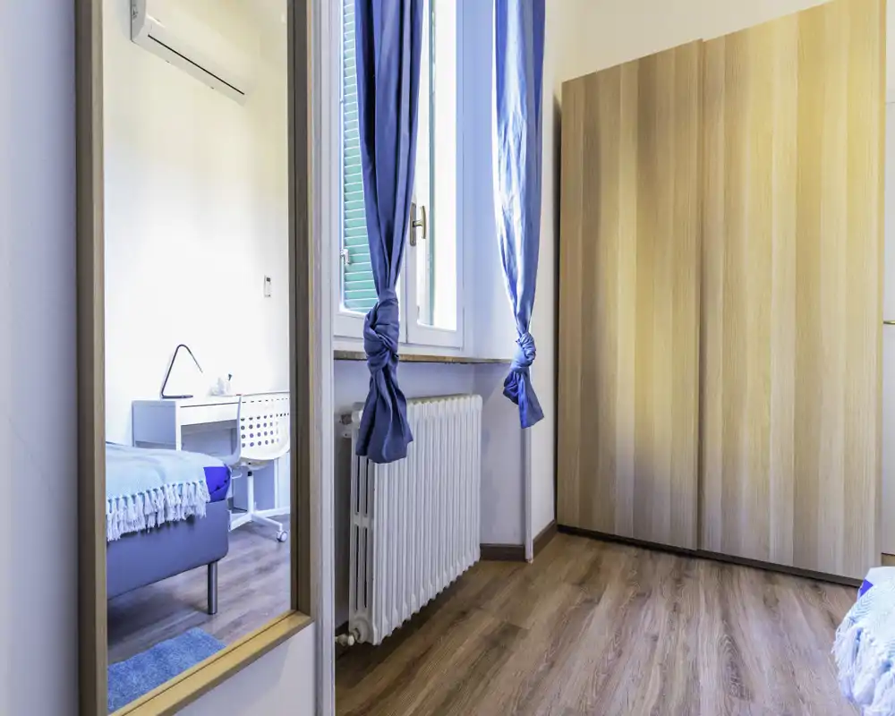 Rincón de dormitorio luminoso y ordenado con toques azules: cama parcialmente visible, escritorio y gran armario de madera junto a una ventana con cortinas azules.
