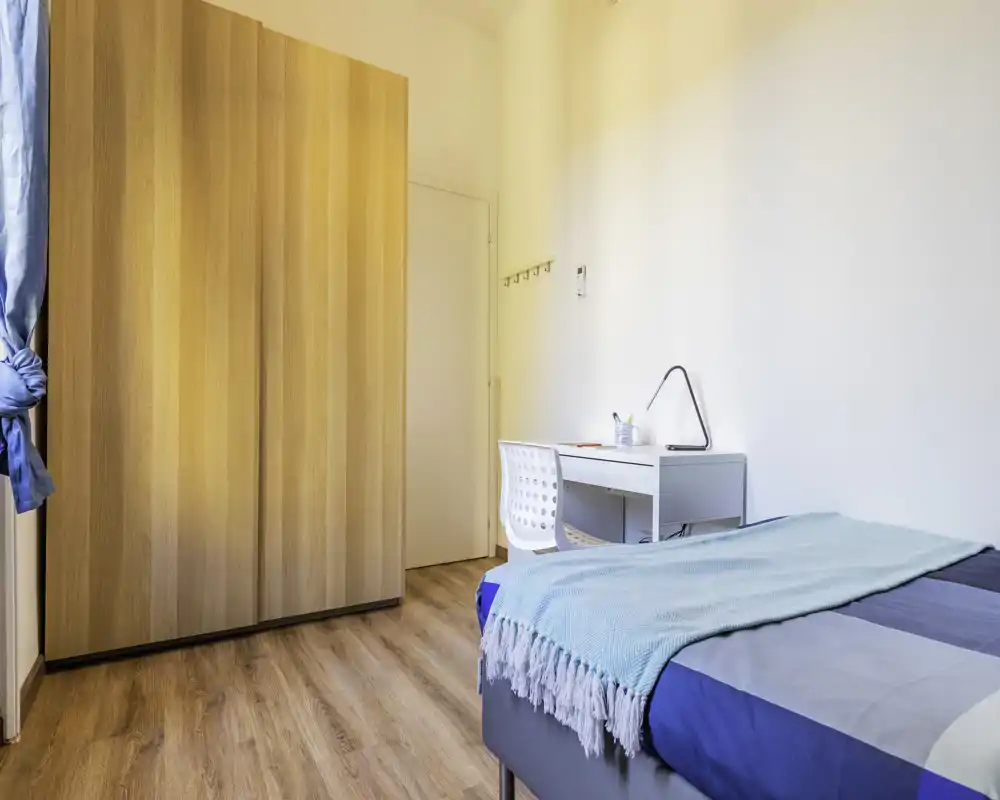 Dormitorio luminoso y ordenado con cama individual y manta azul, escritorio blanco y gran armario de madera. La luz natural y el suelo de madera generan un ambiente tranquilo y acogedor, ideal para alquileres temporales.