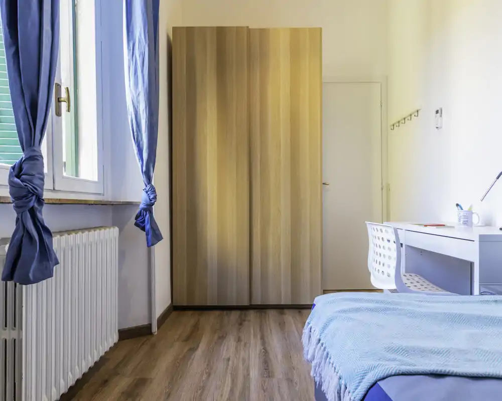 Dormitorio luminoso y ordenado con cama individual con ropa de cama azul, armario de madera, escritorio y luz natural de la ventana.
