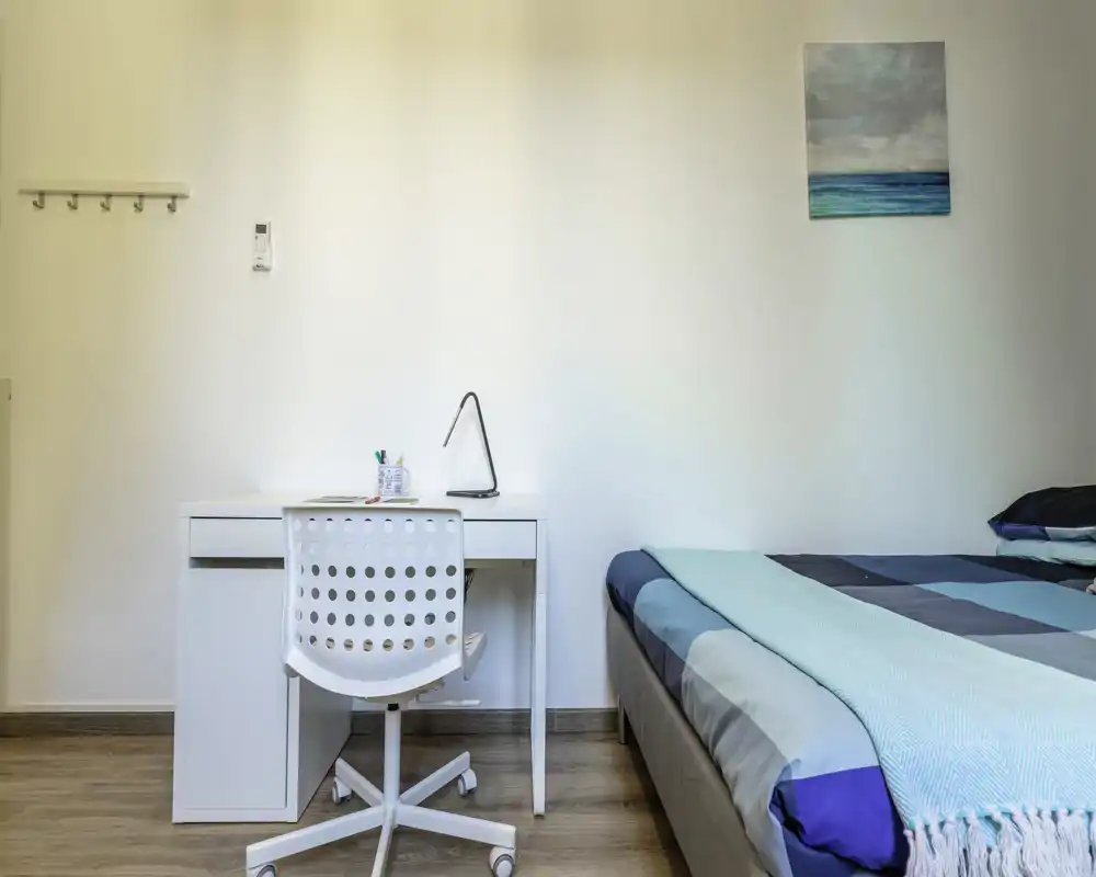 Rincón de dormitorio luminoso y moderno con cama doble con ropa de cama azul-gris y un escritorio blanco sencillo con silla y lámpara — ideal para trabajar y descansar.