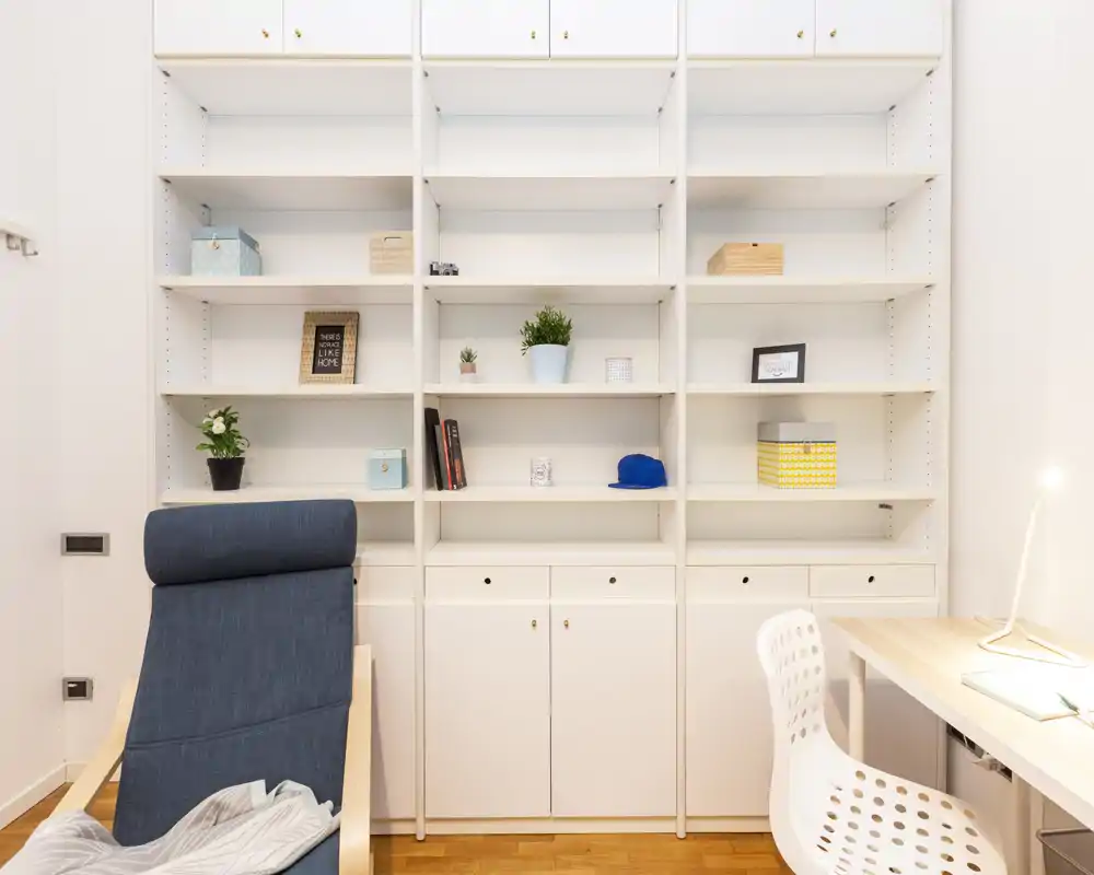 Zona giorno luminosa e moderna con una comoda poltrona blu, scaffalature bianche integrate con piante e libri e una piccola scrivania — ideale per rilassarsi o lavorare da casa.