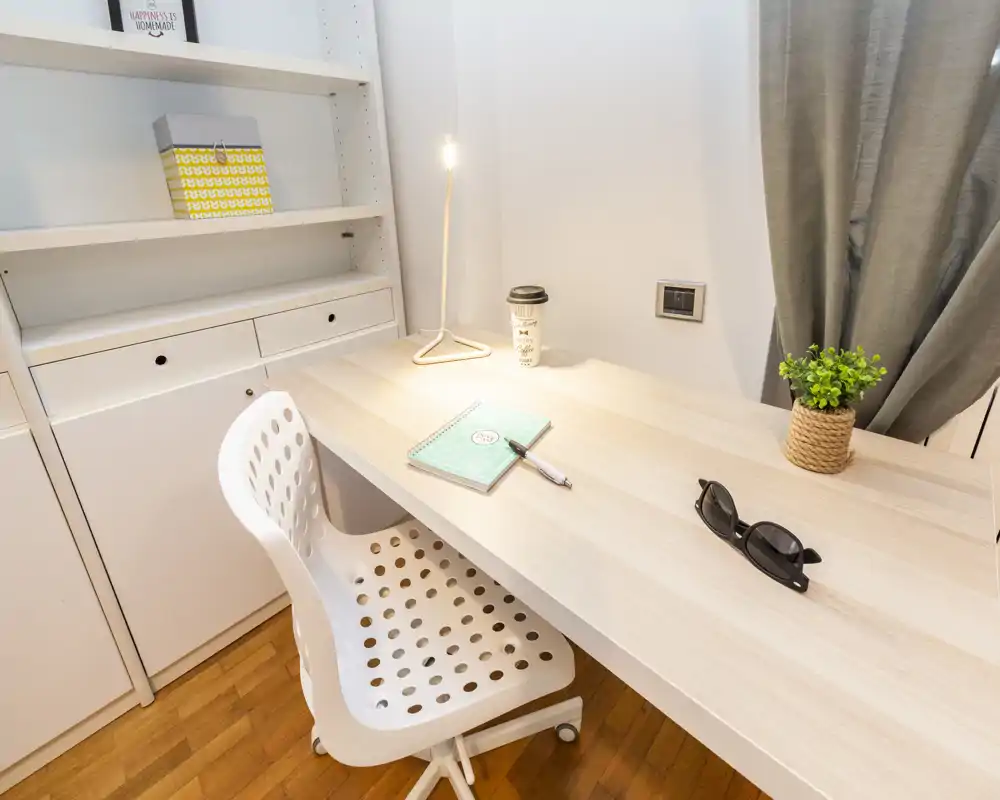 Angolo lavoro luminoso e ordinato con scrivania in legno chiaro, sedia bianca forata, lampada da tavolo, piccola pianta e scaffalatura — perfetto per studiare o lavorare.