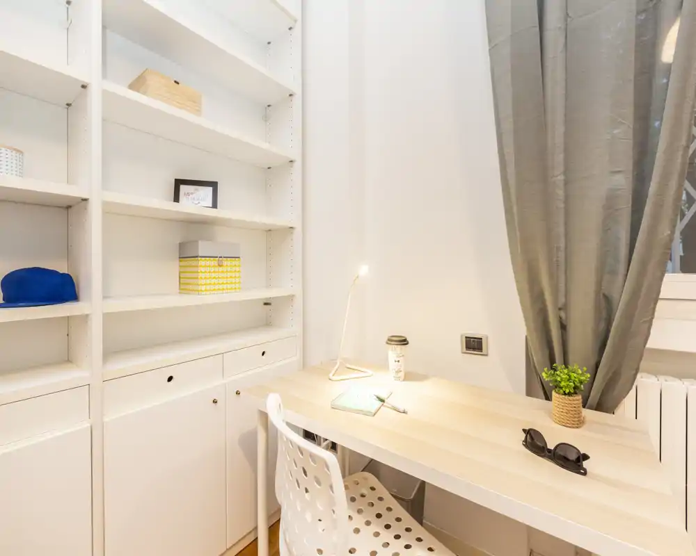 Angolo studio compatto e luminoso con scrivania in legno chiaro, sedia bianca traforata, scaffalatura integrata e lampada da tavolo — perfetto per smart working o studio.