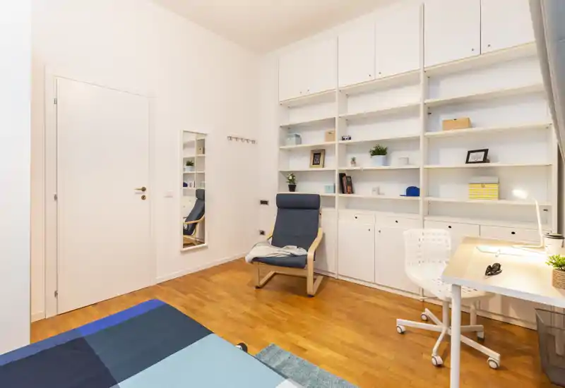 Camera da letto luminosa e moderna con letto ordinato, poltrona per la lettura, scaffalatura a muro e una scrivania — ideale per uno studente o single.