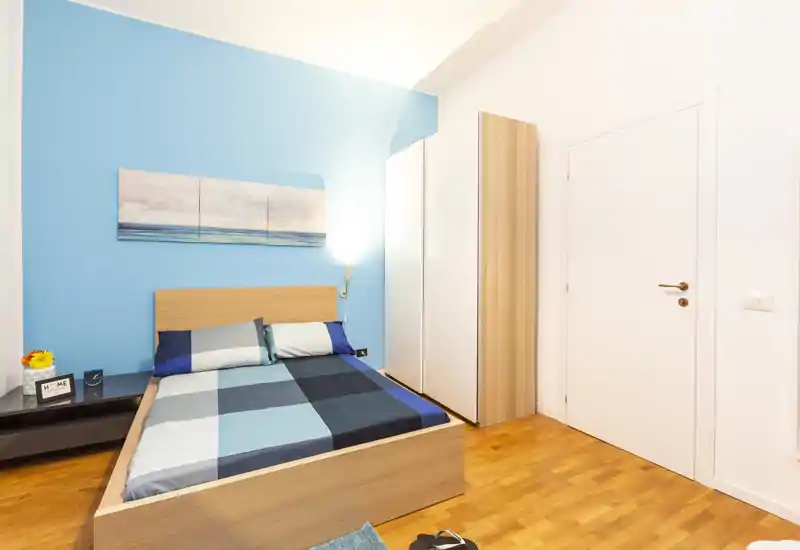 Camera da letto luminosa e moderna con letto matrimoniale, biancheria blu coordinata, comodino e armadio; linee pulite e tinte gradevoli la rendono ideale per annunci.