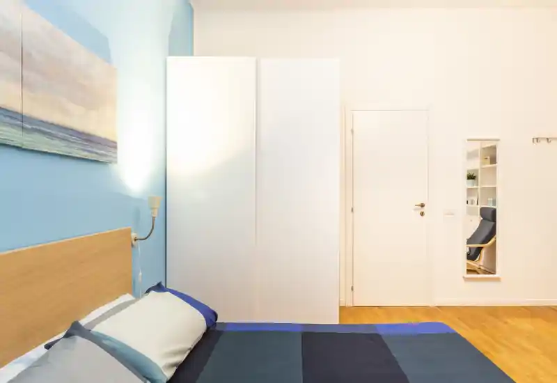 Angolo di camera da letto moderno e luminoso con letto matrimoniale visibile, testiera in legno, lampada da comodino e armadio bianco su parete accentuata azzurra.