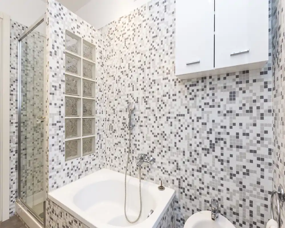 Bagno moderno e luminoso con pareti in mosaico, vasca con doccetta e box doccia separato. Design pulito e neutro, perfetto per annunci di affitto.