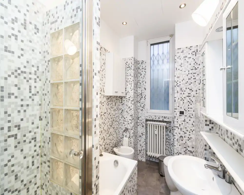 Bagno moderno e luminoso con rivestimenti a mosaico, vasca, bidet e lavabo; finestra satinata per la privacy e faretti a soffitto per una buona illuminazione.
