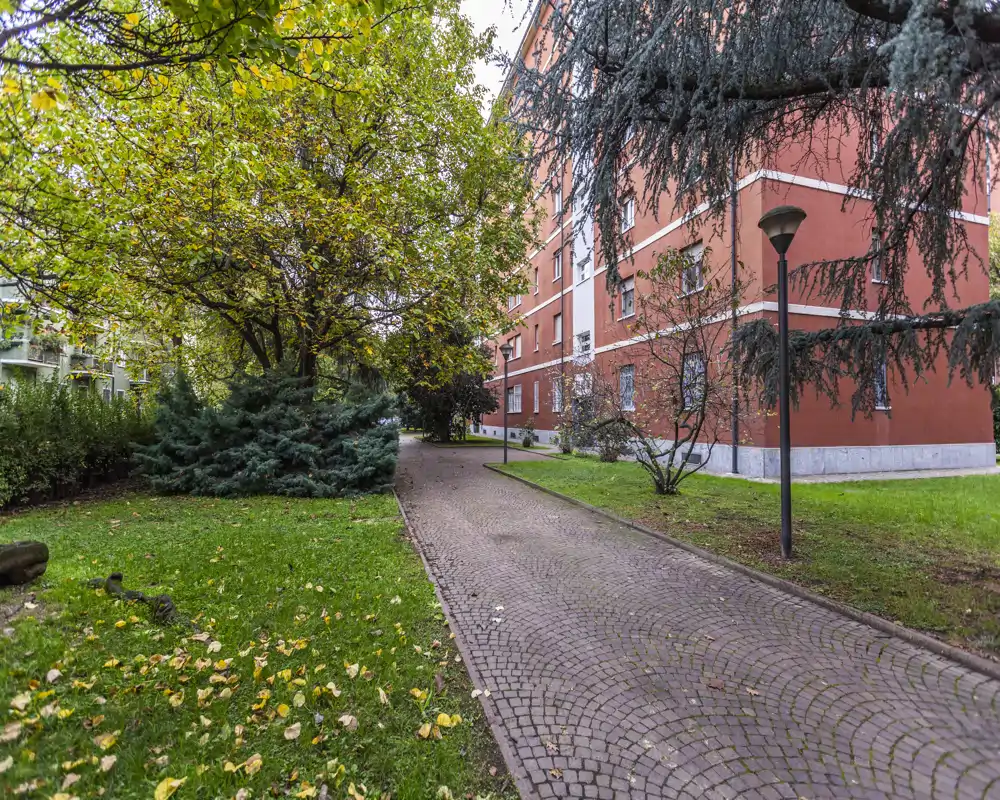 Vialetto lastricato in un cortile con prato e alberi accanto a un edificio residenziale rosso, illuminato da lampioni: area esterna comune tranquilla.