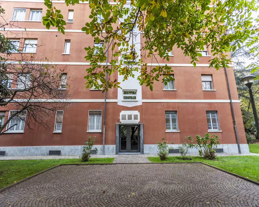 Vista esterna di un edificio residenziale con ingresso, cortile lastricato, aree verdi e rami di alberi sospesi.
