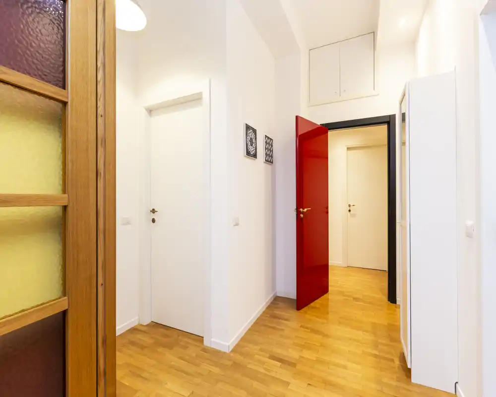 Ingresso luminoso e moderno con pavimento in parquet, pareti bianche e una porta interna rossa che si apre verso l'appartamento.