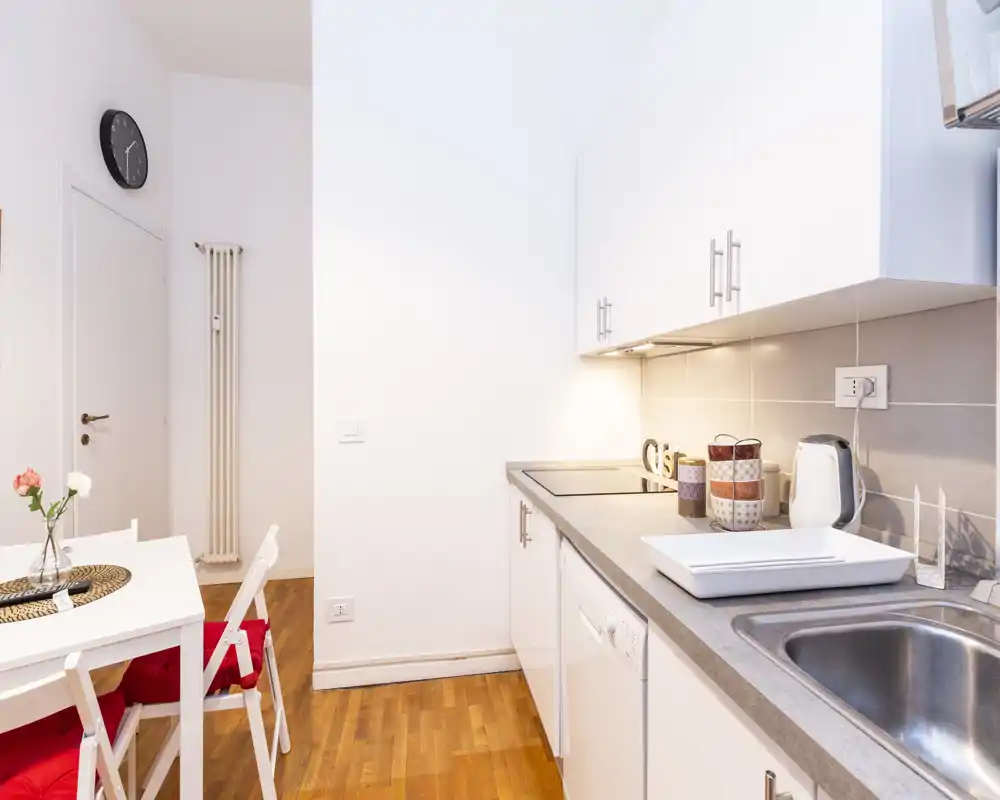 Cucina compatta e luminosa con piano di lavoro moderno, lavello e piano a induzione; piccolo tavolo da pranzo con sedie e decorazioni semplici la rendono pratica e accogliente.