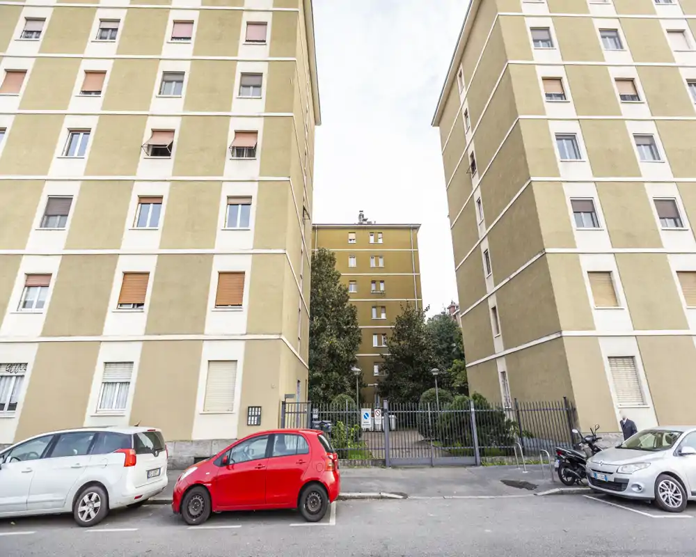 Veduta esterna di palazzi residenziali con auto parcheggiate e un ingresso con cancello che conduce a un cortile. L’inquadratura simmetrica tra i due edifici evidenzia il passaggio d’ingresso e il contesto urbano.