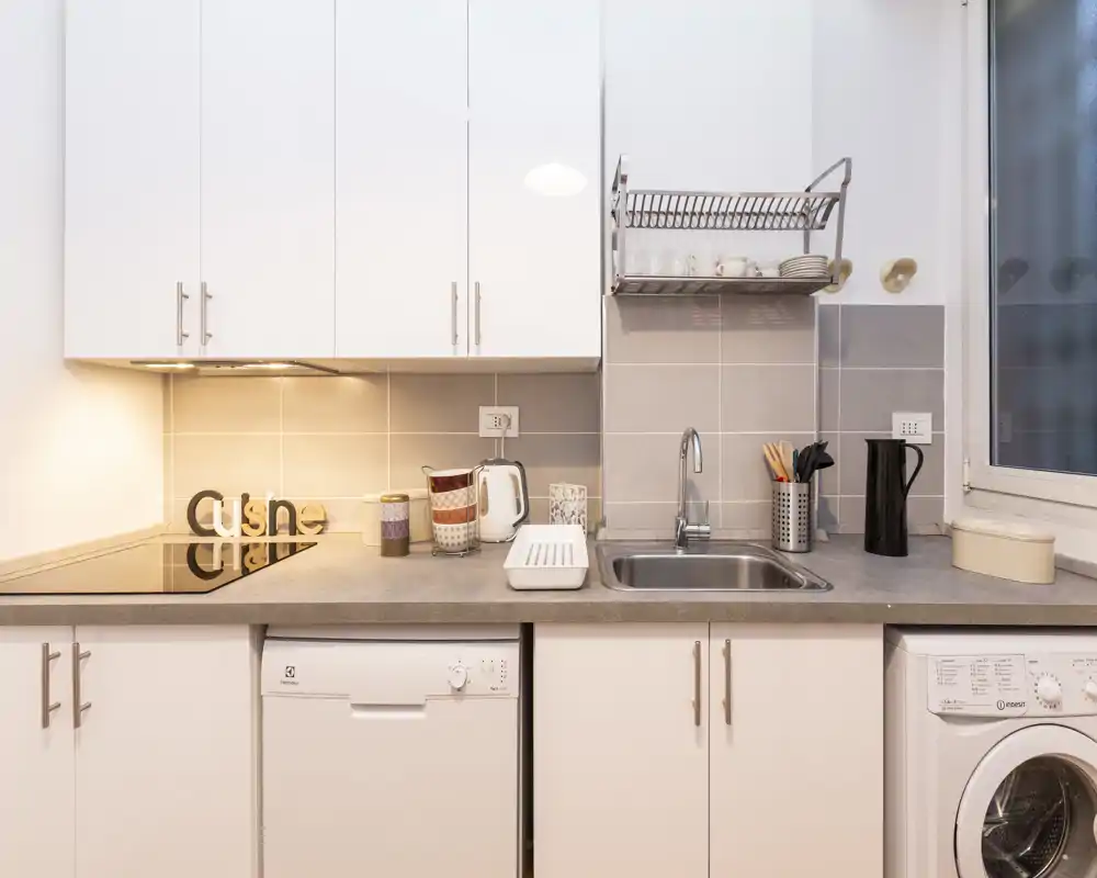 Cucina moderna e luminosa con lavello in acciaio, piano a induzione, lavastoviglie e lavatrice sotto il piano. Piastrelle neutre, mobili bianchi e accessori pratici creano uno spazio pulito e funzionale, ideale per soggiorni brevi.