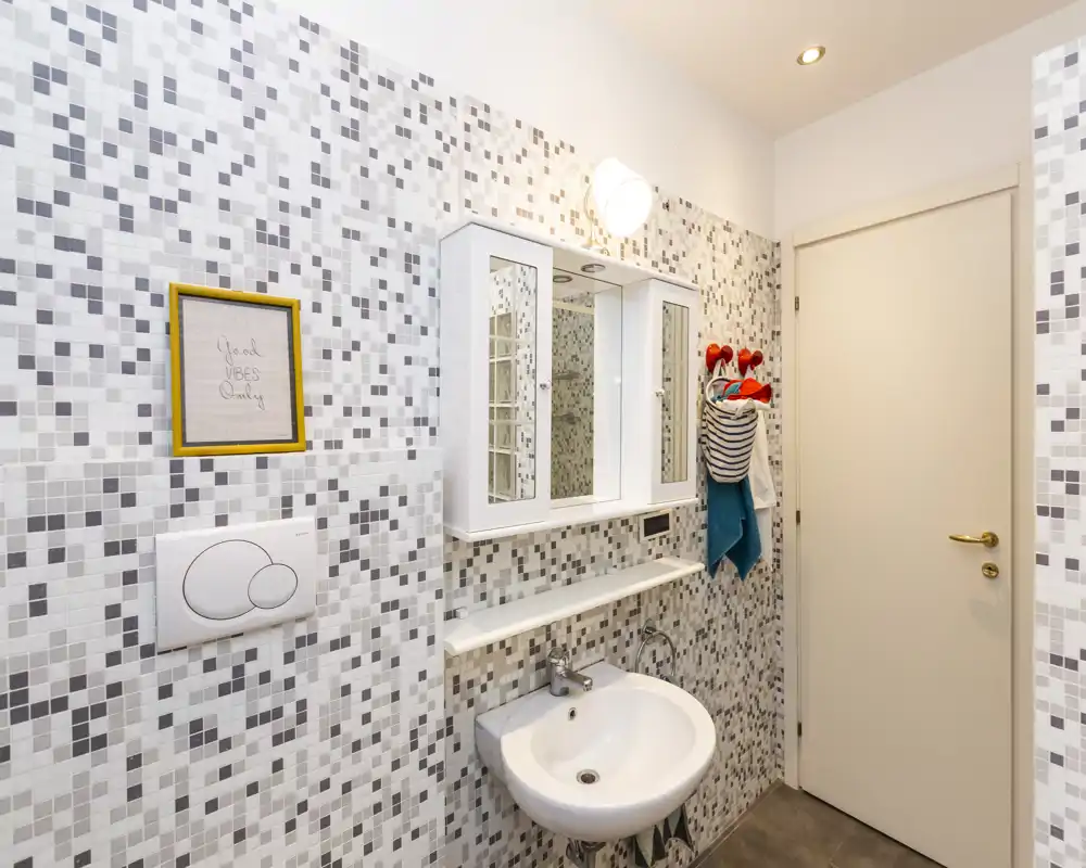 Bagno moderno di piccole dimensioni con rivestimento a mosaico, lavabo a parete e mobile specchiera. Design luminoso e pulito, layout compatto ideale per un appartamento in città.