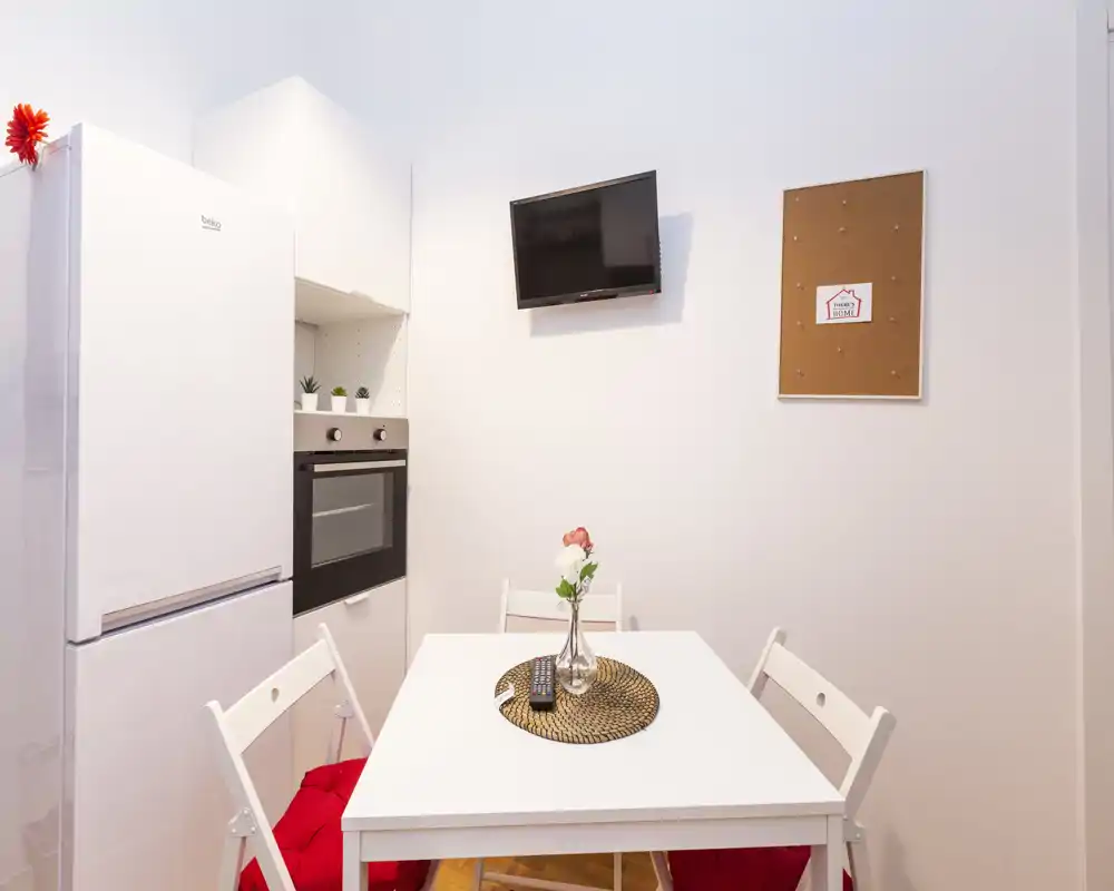 Angolo cucina moderno e compatto con tavolo da pranzo bianco e sedie, forno incassato, frigorifero e una piccola TV a muro. Il tavolo è decorato con un sottopiatto intrecciato e un vaso con fiori, creando un angolo pranzo ordinato e luminoso.