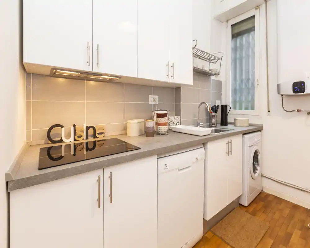 Cucina moderna e compatta con pensili bianchi, piano ad induzione, lavastoviglie integrata e lavatrice; piano di lavoro grigio chiaro e pavimento in legno caldo per un ambiente pulito e accogliente.