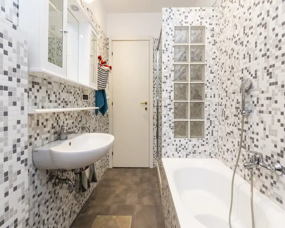 Bagno moderno e pulito con piastrelle a mosaico, lavabo a parete, vasca con doccia a mano e blocchi di vetro satinato per la privacy.