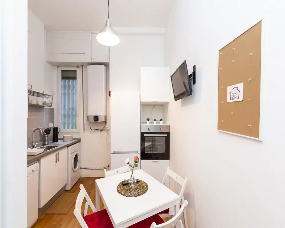 Cucina compatta e moderna con elettrodomestici integrati, piccolo tavolo bianco con sedie, TV a parete e stile luminoso e pulito — ideale per un monolocale cittadino.