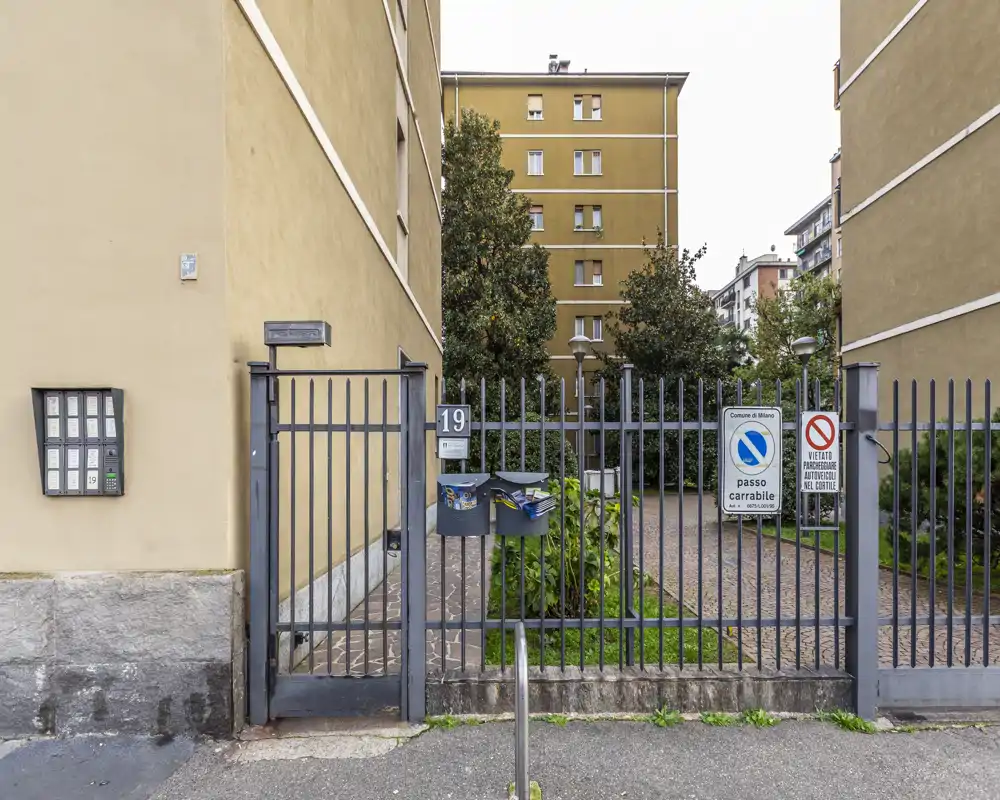 Ingresso condominiale con cancellata, citofoni e cancello pedonale che si apre su un cortile con verde e l'edificio residenziale sullo sfondo.