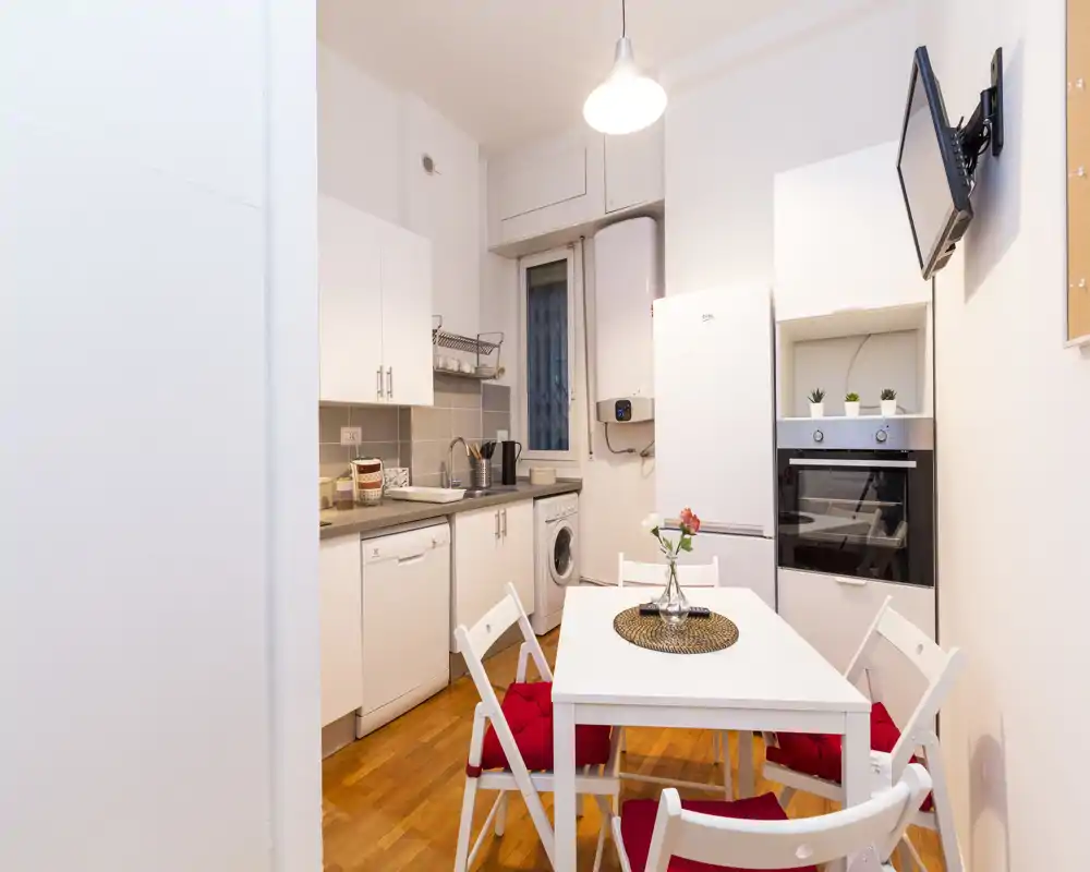 Cucina compatta e luminosa con elettrodomestici integrati, un piccolo tavolo da pranzo e quattro sedie con cuscini rossi — ideale per un appartamento cittadino accogliente.
