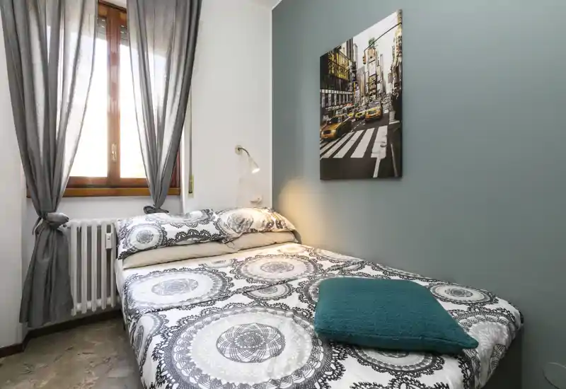 Camera da letto compatta e luminosa con letto matrimoniale, biancheria a motivi, cuscino turchese, lampada a parete e finestra con tende grigie.