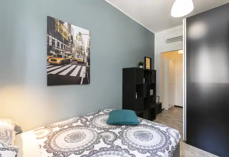 Camera da letto luminosa e ben arredata con copriletto decorato, cuscino turchese come accento e quadro urbano; armadio e scaffale offrono spazio e ordine.