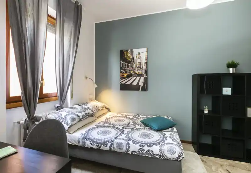 Camera da letto luminosa e ben arredata con letto matrimoniale, biancheria a motivi e cuscino decorativo vicino alla finestra con tende. La stanza ha una parete accent color verde acqua, lampada da comodino e scaffale, ideale per affitti brevi.