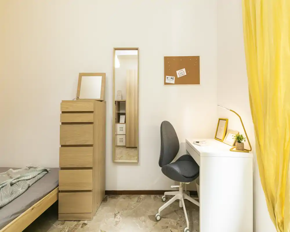 Coin chambre lumineux et minimaliste avec le bord d’un lit simple visible, une commode en bois haute, un miroir en pied et un bureau blanc soigné avec chaise et lampe.