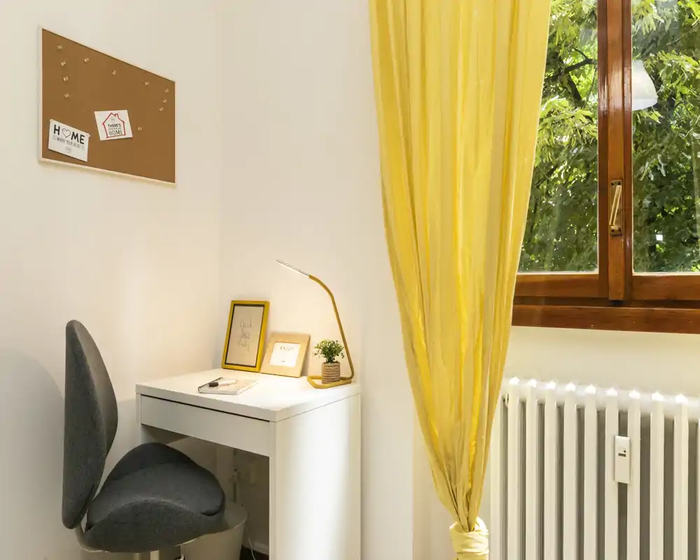 Coin salon lumineux et compact avec un petit bureau blanc, une chaise grise moderne et un rideau jaune vif encadrant une grande fenêtre — idéal pour lire ou travailler à la lumière naturelle.