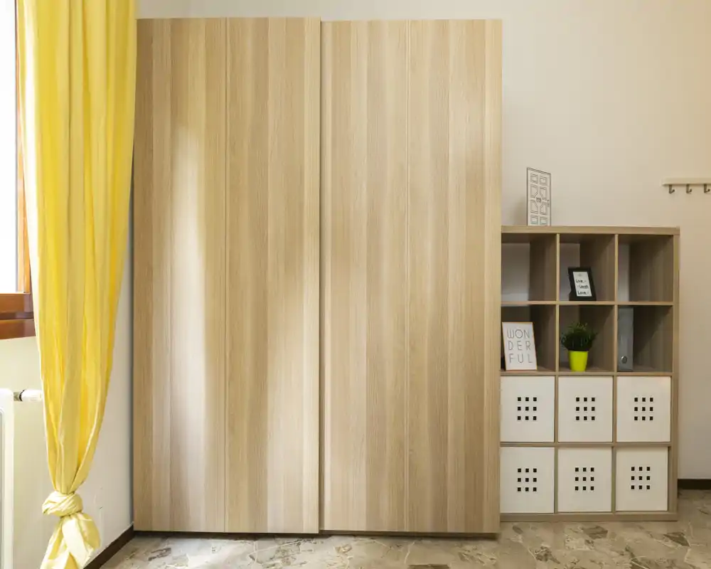 Vue de rangement de chambre lumineuse avec une grande armoire en bois clair et des cubes de rangement décorés, éclairée par la lumière naturelle d'une fenêtre proche.