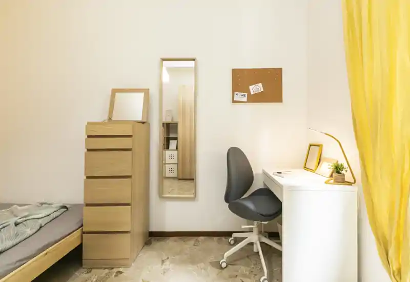 Coin chambre lumineux et minimaliste avec le bord d’un lit simple visible, une commode en bois haute, un miroir en pied et un bureau blanc soigné avec chaise et lampe.