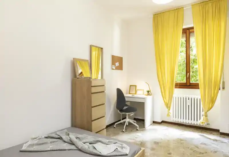 Chambre lumineuse et sobre avec un lit simple, une commode en bois, un bureau d'étude et une grande fenêtre avec des rideaux jaunes vifs laissant entrer la lumière naturelle.