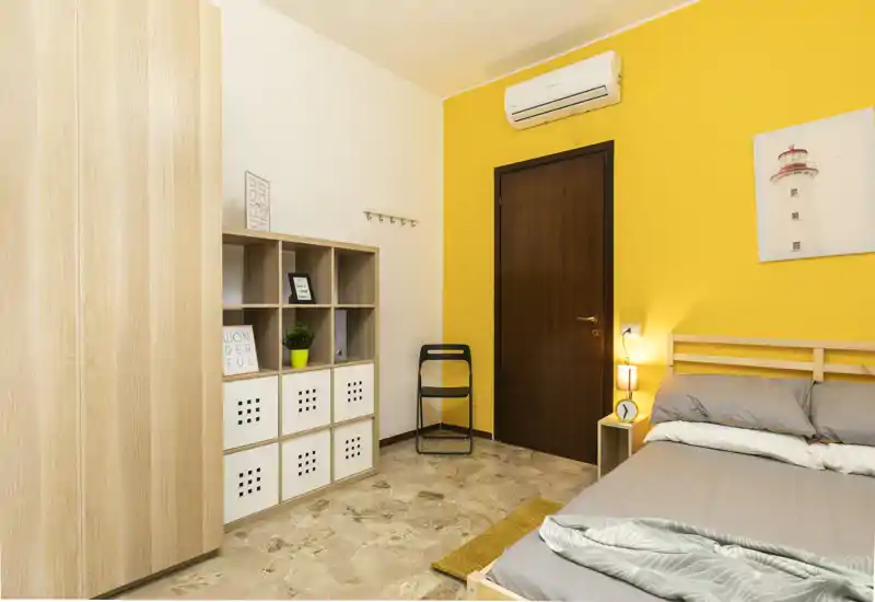 Chambre moderne et lumineuse avec cadre de lit en bois, literie grise et mur d’accent jaune chaleureux. La pièce dispose d’une armoire, d’étagères cubiques, d’une lampe de chevet et d’une climatisation — idéale pour un séjour confortable.