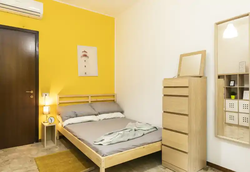 Chambre lumineuse et soignée avec un lit double en bois, une lampe de chevet et une grande commode. Le mur jaune vif et le linge de lit apportent une atmosphère chaleureuse et accueillante, idéale pour une annonce.