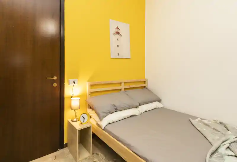 Chambre compacte et lumineuse avec un cadre de lit en bois, une literie grise neutre et un mur d'accent jaune chaleureux. La pièce est bien éclairée et simplement aménagée, parfaite pour une personne ou des séjours courts.