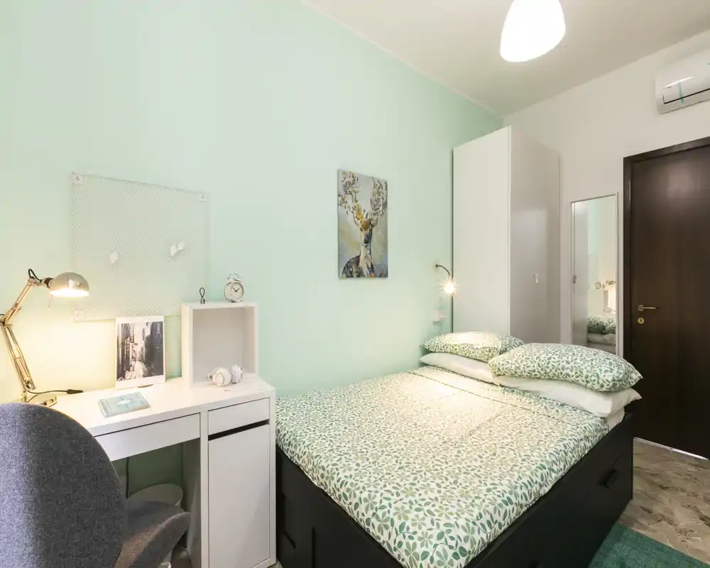 Dormitorio compacto y luminoso con cama doble bien hecha, ropa de cama verde a juego, un pequeño escritorio blanco con lámpara y un armario — ideal para una persona o pareja que busca un espacio acogedor y bien iluminado.