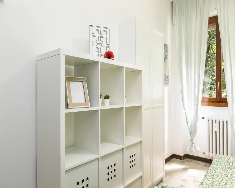 Rincón de dormitorio luminoso y minimalista con estantería modular blanca, un armario alto y una ventana que aporta luz natural; se aprecia una pequeña porción de la cama con funda estampada.